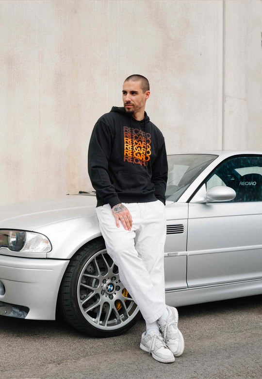 Ein Mann, der einen „Recaro_Merch_Daniel“-Hoodie und eine schwarz-weiße Hose trägt, lehnt an einer silbernen BMW vor einer beigen Wand und nimmt mit den Händen in den Taschen eine Retro-Pose ein. Sein kurzes Haar und sein Bart unterstreichen den Unisex-Stil des Hoodies.