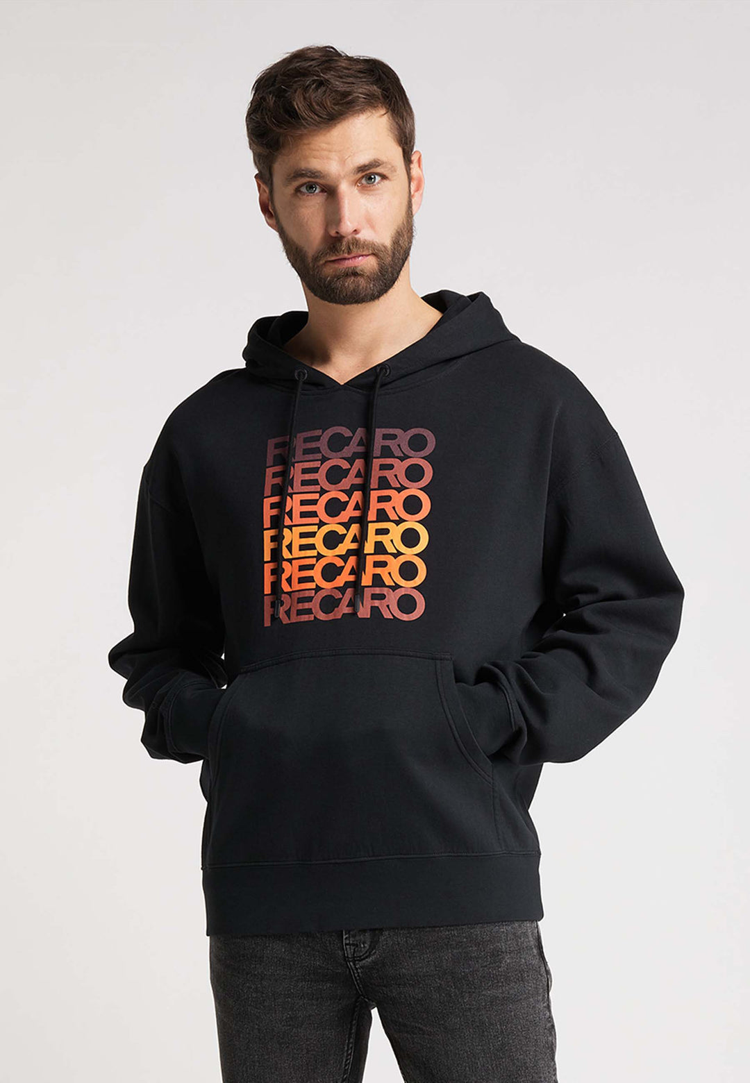 Ein bärtiger Mann steht vor der Kamera in einem Recaro_Merch_Daniel Hoodie Spektrum mit einem Retro-Design mit "RECARO" in rotem, orangefarbenem und gelbem Farbverlauf, gepaart mit einer dunklen Jeans vor einem schlichten hellen Hintergrund.