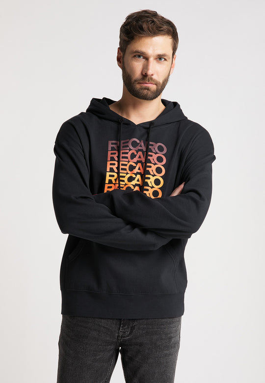 Ein bärtiger Mann steht mit verschränkten Armen da und trägt einen schwarzen Recaro_Merch_Daniel Hoodie Spektrum mit orangefarbenen und gelben Retro-Logos vor einem schlichten weißen Hintergrund.