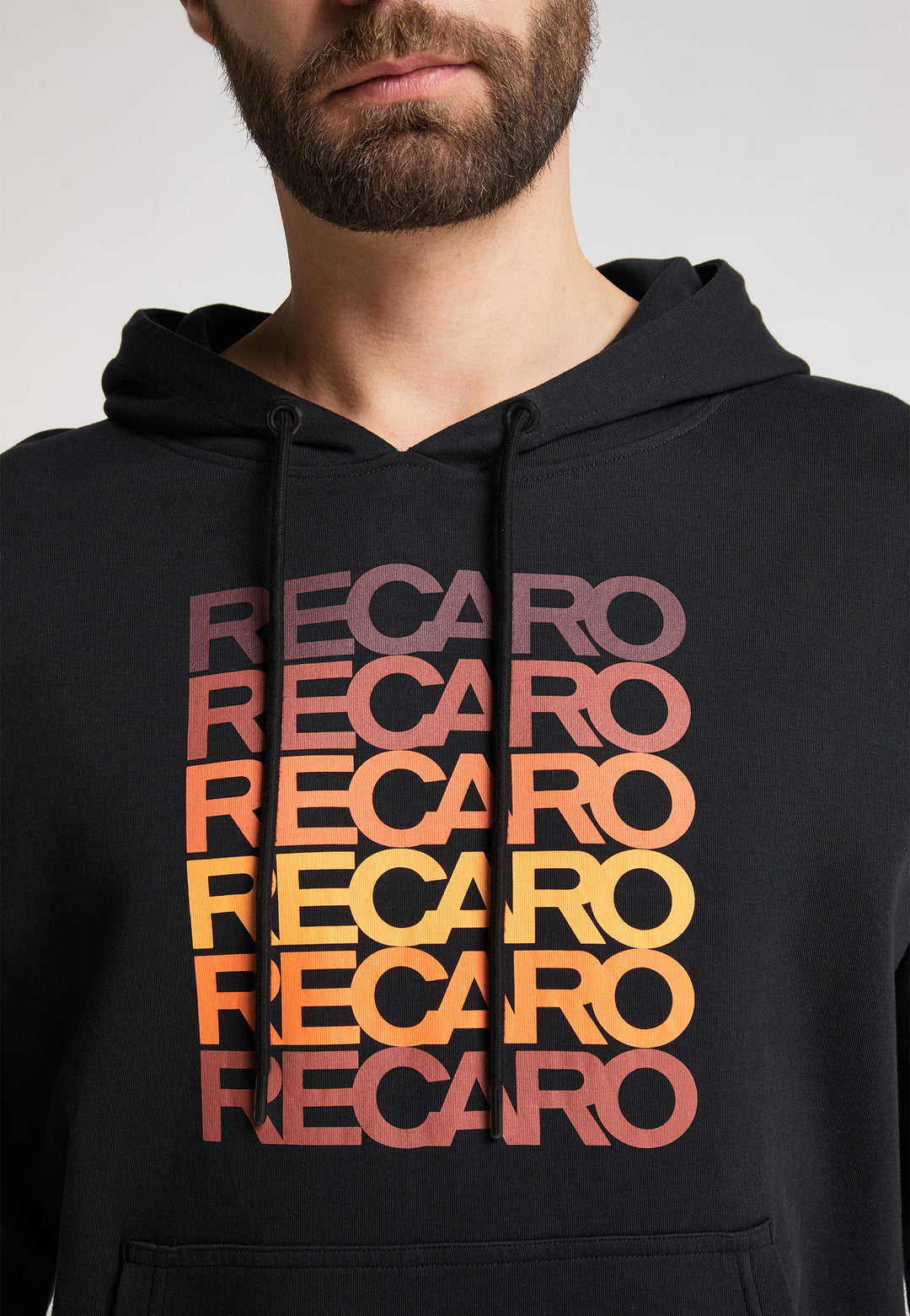 Ein Mann mit Bart trägt den Recaro_Merch_Daniel Hoodie Spektrum mit dem fünfmal wiederholten Schriftzug RECARO" in einem Retro-Rot-Gelb-Verlauf. Nur sein unteres Gesicht und sein oberer Oberkörper sind sichtbar.