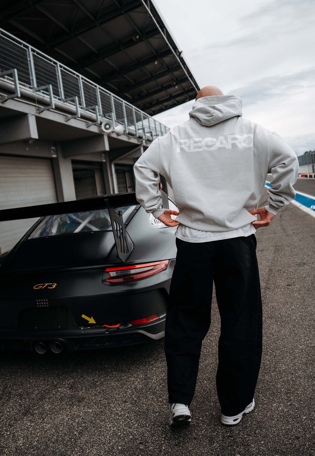 In einem RECARO Shop Hoodie RECARO Heavy Cotton steht jemand neben einem schwarzen Porsche GT3 an der Rennstrecke, mit einem auffälligen Logo auf dem Rücken, mit Blick auf die Boxengasse und die Garage.