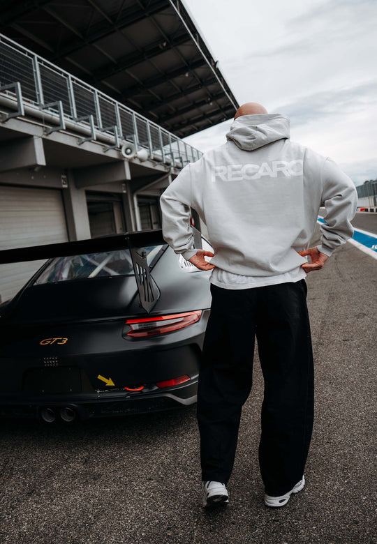 In einem RECARO Shop Hoodie RECARO Heavy Cotton steht jemand neben einem schwarzen Porsche GT3 an der Rennstrecke, mit einem auffälligen Logo auf dem Rücken, mit Blick auf die Boxengasse und die Garage.