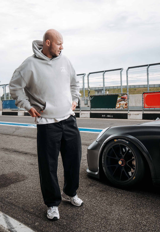 Ein Mann, der den RECARO Shop's Hoodie RECARO Heavy Cotton und eine schwarze Hose trägt, steht neben einem schwarzen Sportwagen auf einer Rennstrecke. Er blickt auf das Auto, weiße Turnschuhe und die Boxenmauer sind zu sehen, dazu ein dezentes RECARO Logo im Hintergrund.