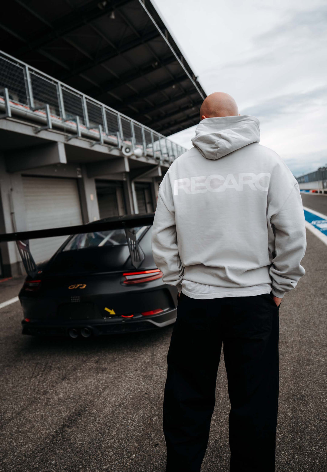 Eine Person, die den RECARO Shop's Hoodie RECARO Heavy Cotton trägt, steht auf einer Rennstrecke neben einem schwarzen Porsche GT3, mit Garagengebäuden im Hintergrund.