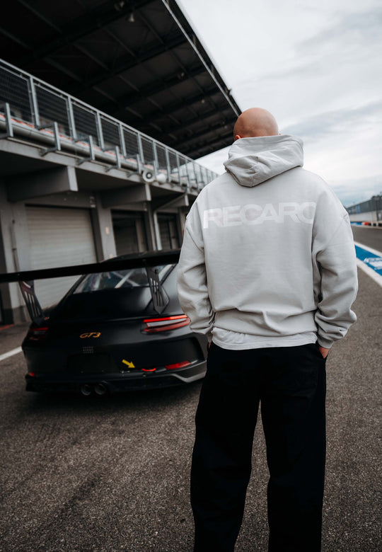 Eine Person, die den RECARO Shop's Hoodie RECARO Heavy Cotton trägt, steht auf einer Rennstrecke neben einem schwarzen Porsche GT3, mit Garagengebäuden im Hintergrund.