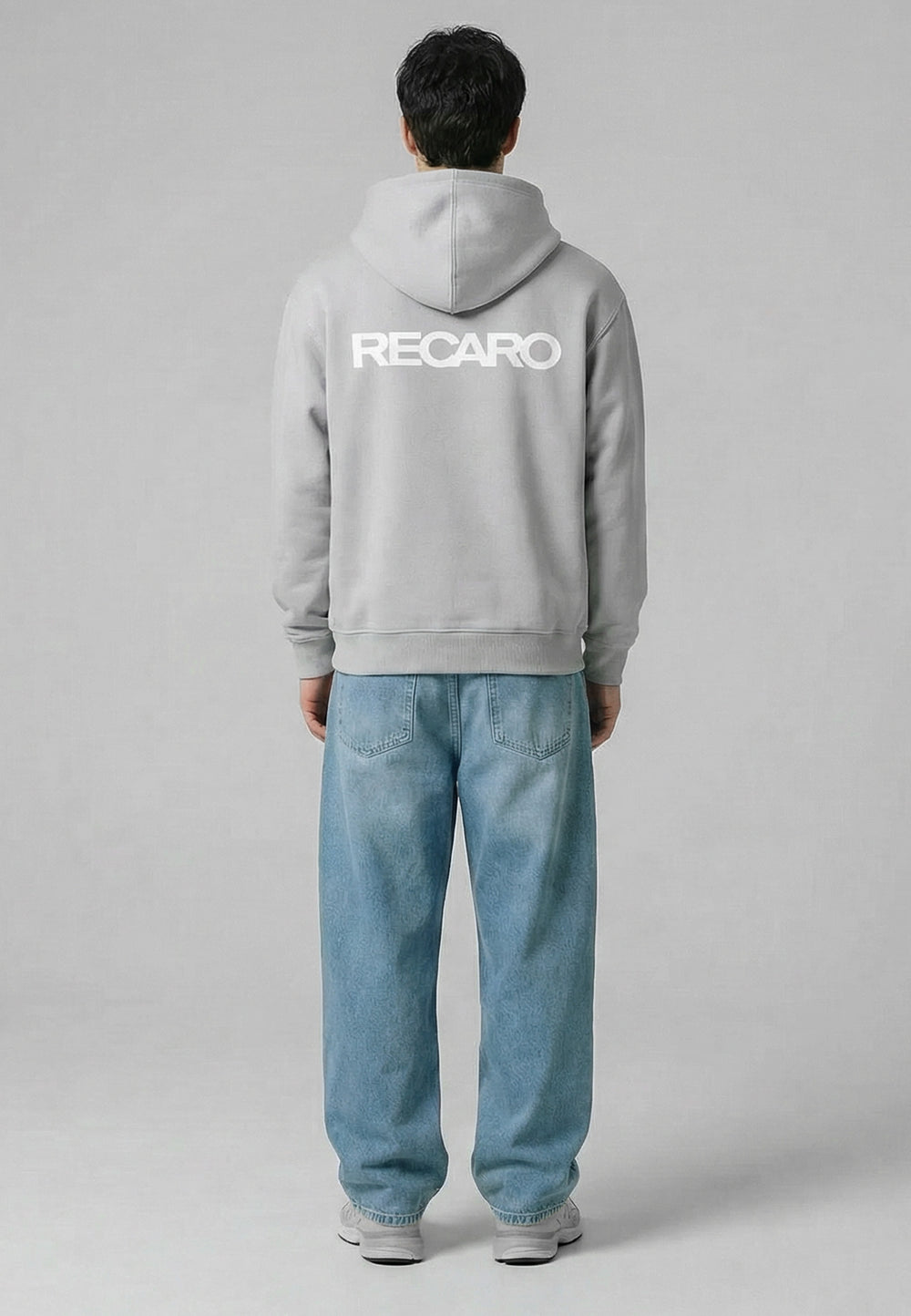 Eine Person steht mit dem Gesicht abgewandt, trägt einen hellgrauen Kapuzenpullover RECARO Heavy Cotton aus dem RECARO Shop mit dem Logo in großen weißen Buchstaben auf dem Rücken, dazu weite blaue Jeans und weiße Turnschuhe, vor einem schlichten grauen Hintergrund.