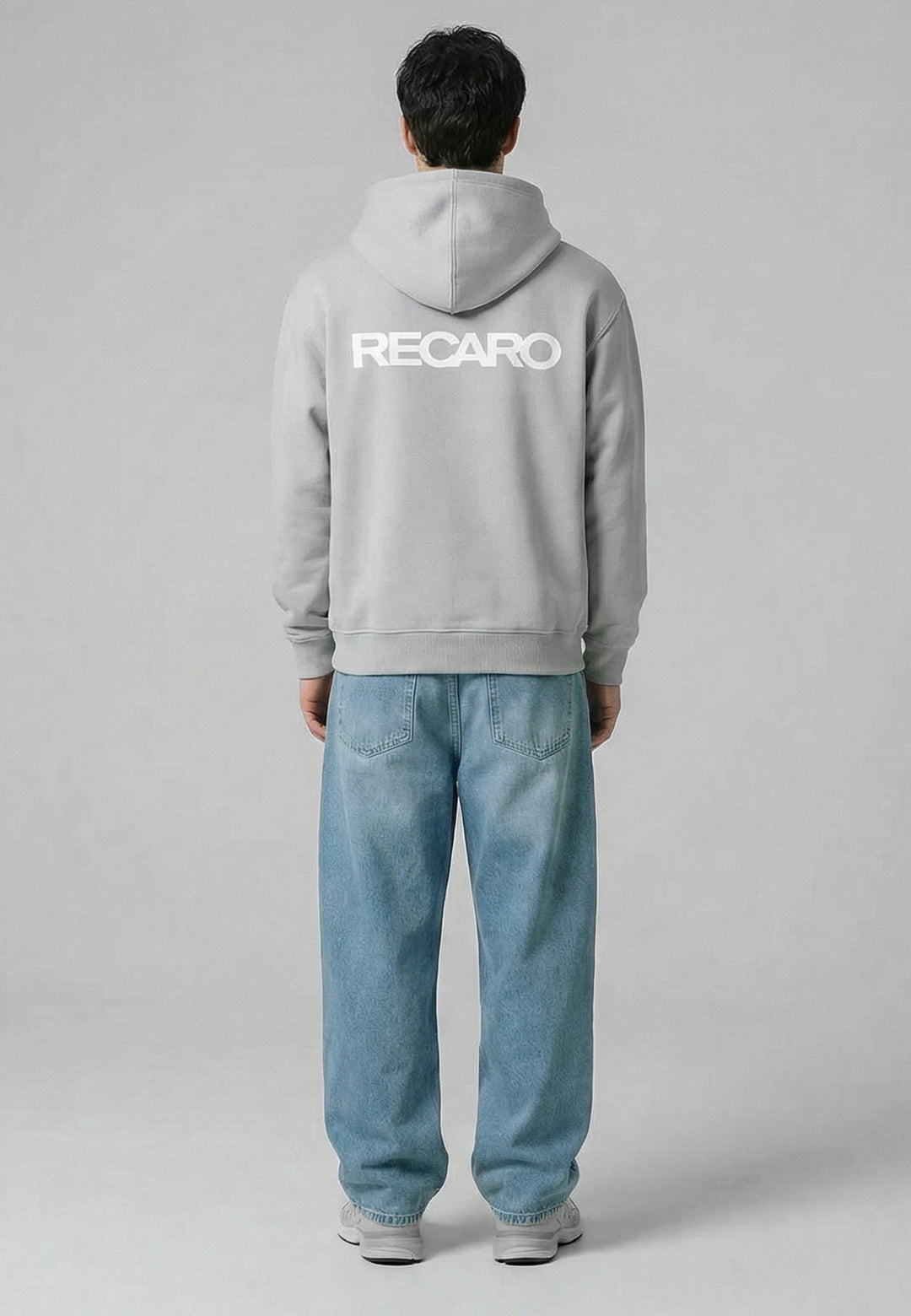 Eine Person steht mit dem Gesicht abgewandt, trägt einen hellgrauen Kapuzenpullover RECARO Heavy Cotton aus dem RECARO Shop mit dem Logo in großen weißen Buchstaben auf dem Rücken, dazu weite blaue Jeans und weiße Turnschuhe, vor einem schlichten grauen Hintergrund.