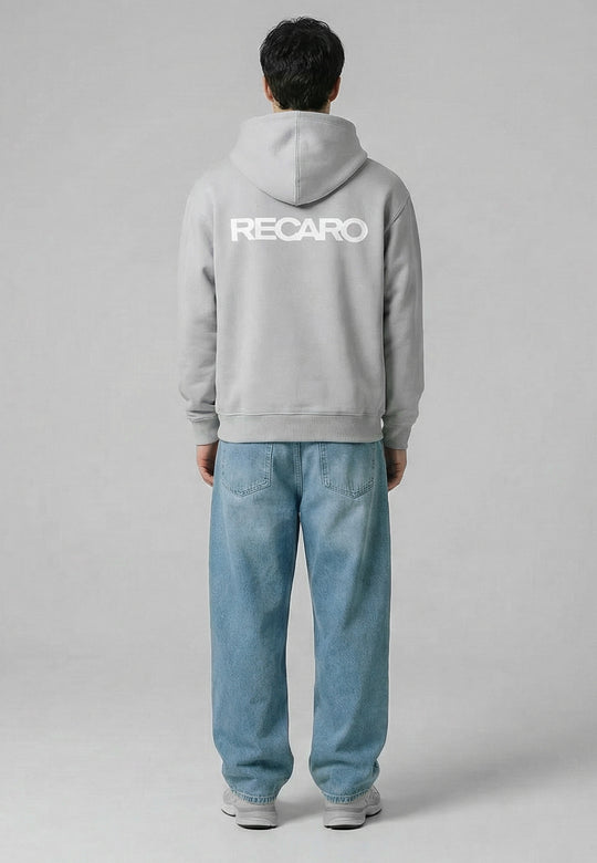 Eine Person steht mit dem Gesicht abgewandt, trägt einen hellgrauen Kapuzenpullover RECARO Heavy Cotton aus dem RECARO Shop mit dem Logo in großen weißen Buchstaben auf dem Rücken, dazu weite blaue Jeans und weiße Turnschuhe, vor einem schlichten grauen Hintergrund.