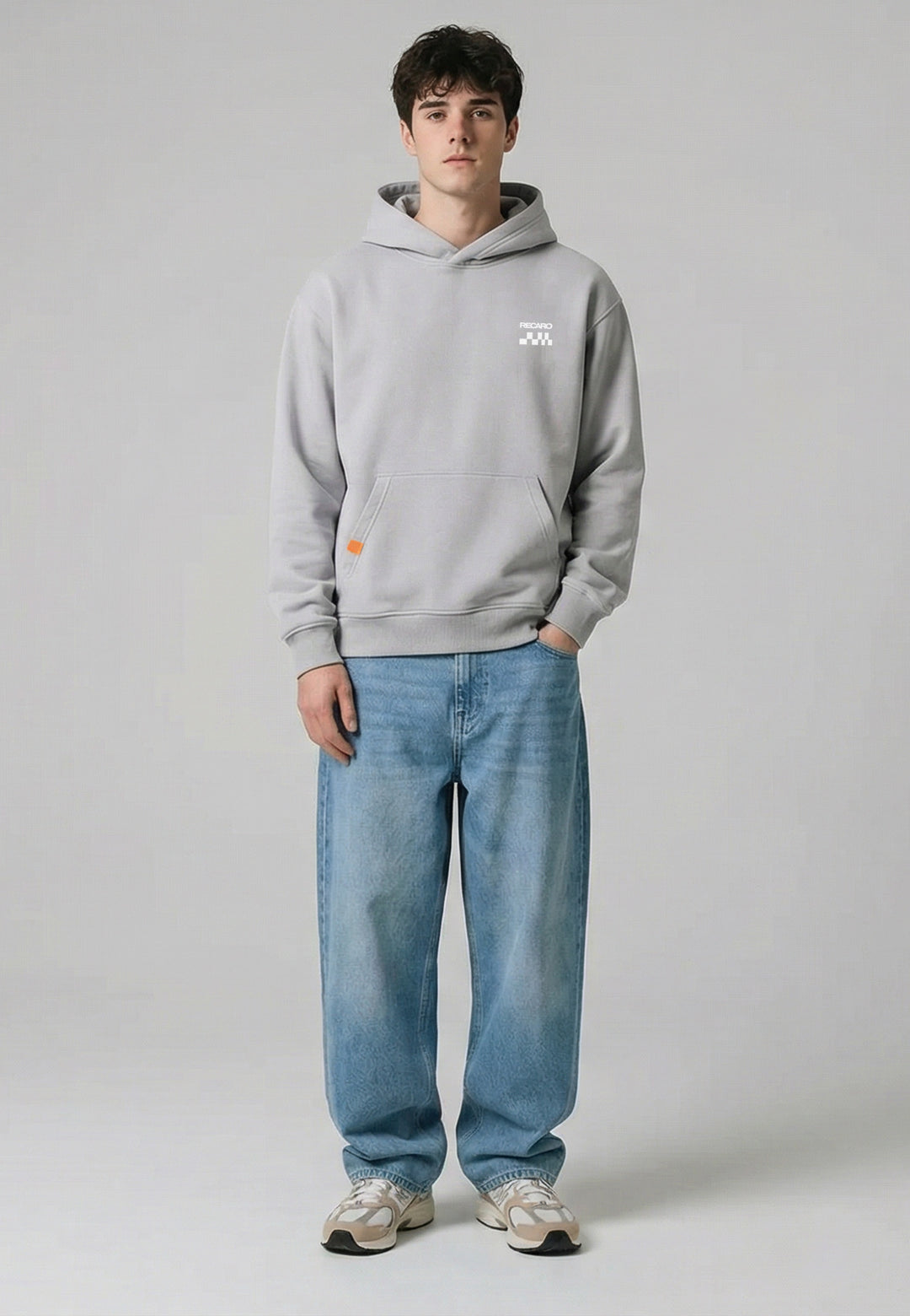 Ein junger Mann steht mit dem Gesicht nach vorne, trägt einen RECARO Shop Hoodie RECARO Heavy Cotton, eine locker sitzende blaue Jeans und beigefarbene Turnschuhe vor einem schlichten hellgrauen Hintergrund.