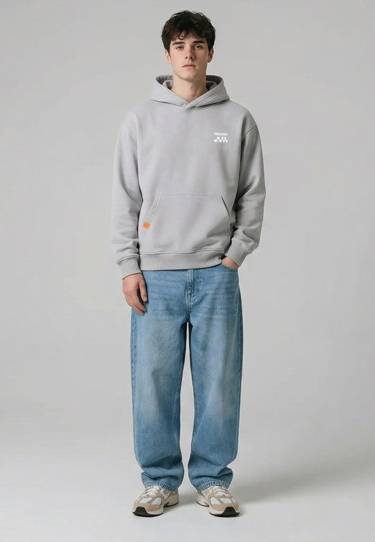 Ein junger Mann steht mit dem Gesicht nach vorne, trägt einen RECARO Shop Hoodie RECARO Heavy Cotton, eine locker sitzende blaue Jeans und beigefarbene Turnschuhe vor einem schlichten hellgrauen Hintergrund.