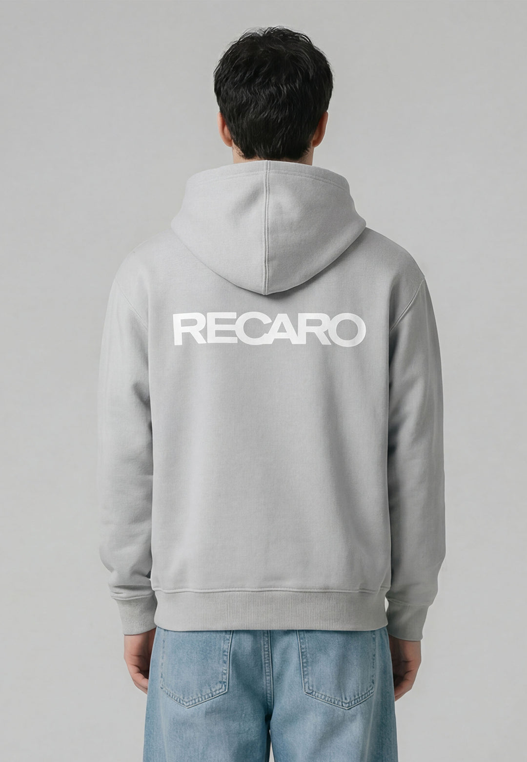 Eine Person blickt vor einem unifarbenen Hintergrund von der Kamera weg. Sie trägt den übergroßen hellgrauen Hoodie RECARO Heavy Cotton von RECARO Shop aus ringgesponnener Baumwolle mit einem auffälligen RECARO Logo auf dem Rücken, gepaart mit einer hellblauen Jeans.