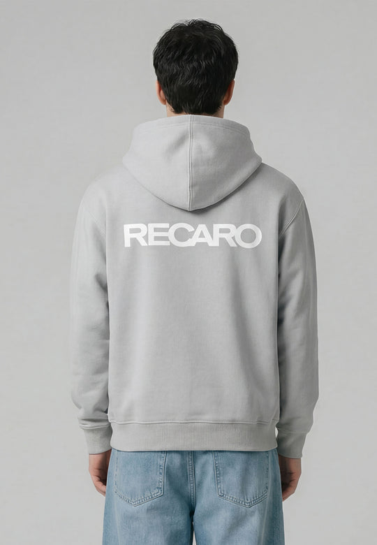 Eine Person blickt vor einem unifarbenen Hintergrund von der Kamera weg. Sie trägt den übergroßen hellgrauen Hoodie RECARO Heavy Cotton von RECARO Shop aus ringgesponnener Baumwolle mit einem auffälligen RECARO Logo auf dem Rücken, gepaart mit einer hellblauen Jeans.