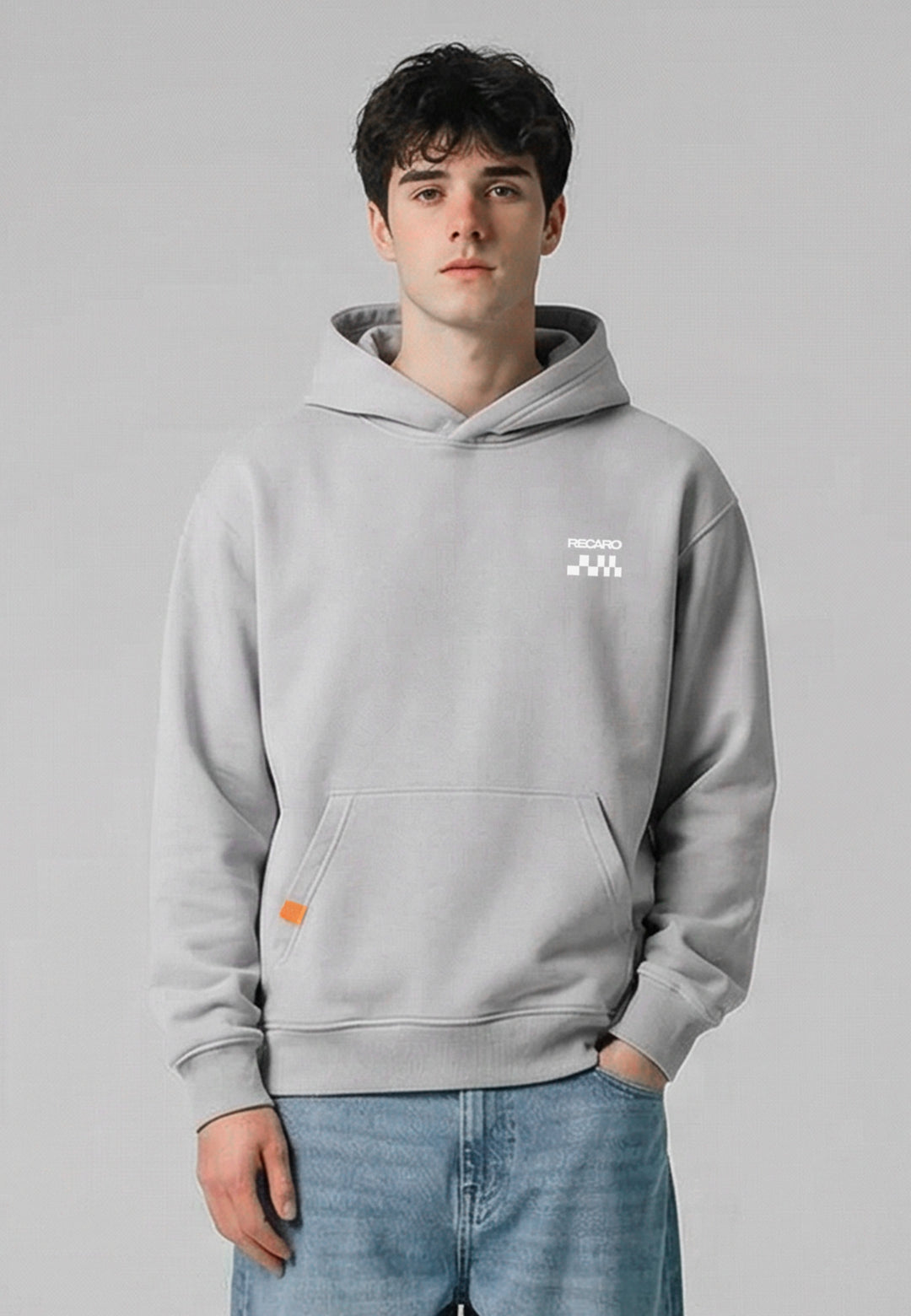 Ein junger Mann mit kurzen dunklen Haaren trägt den übergroßen hellgrauen Hoodie RECARO Heavy Cotton aus dem RECARO Shop und eine blaue Jeans vor einem schlichten hellgrauen Hintergrund. Eine Hand steckt in seiner Hosentasche, während er mit neutralem Blick direkt in die Kamera schaut.