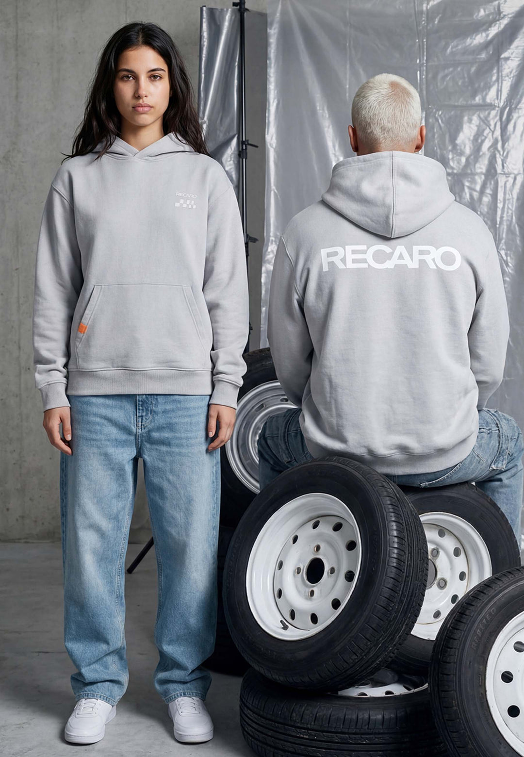 Zwei Personen in grauen Kapuzenpullis und blauen Jeans posieren in einer Garage. Einer sitzt auf Reifen und trägt den RECARO Shop's Hoodie RECARO Heavy Cotton, umgeben von gestapelten Autoreifen.