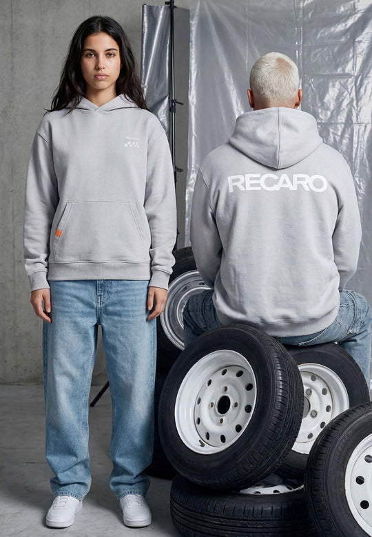 Zwei Personen in grauen Kapuzenpullis und blauen Jeans posieren in einer Garage. Einer sitzt auf Reifen und trägt den RECARO Shop's Hoodie RECARO Heavy Cotton, umgeben von gestapelten Autoreifen.