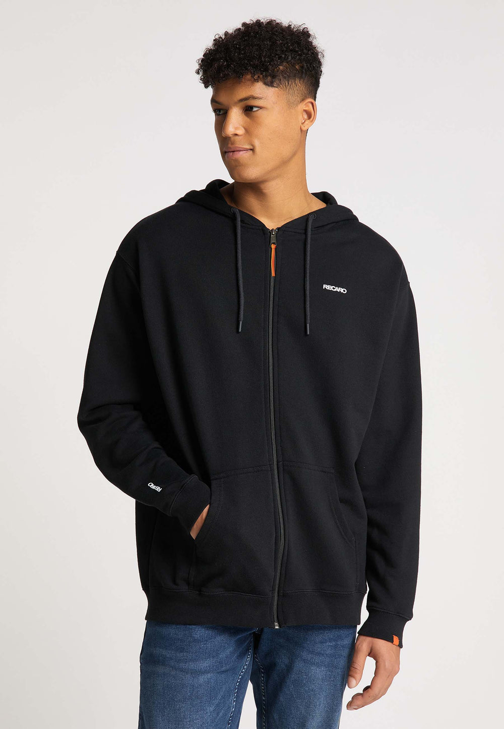 Ein junger Mann steht mit einer Hand in der Tasche da und blickt zur Seite. Er trägt Bluejeans und eine schwarze Hoodiejacke „Originals“ von Recaro_Merch_Daniel – eine locker sitzende Unisex-Jacke mit Reißverschluss, Vordertaschen und kleinem weißen Text.