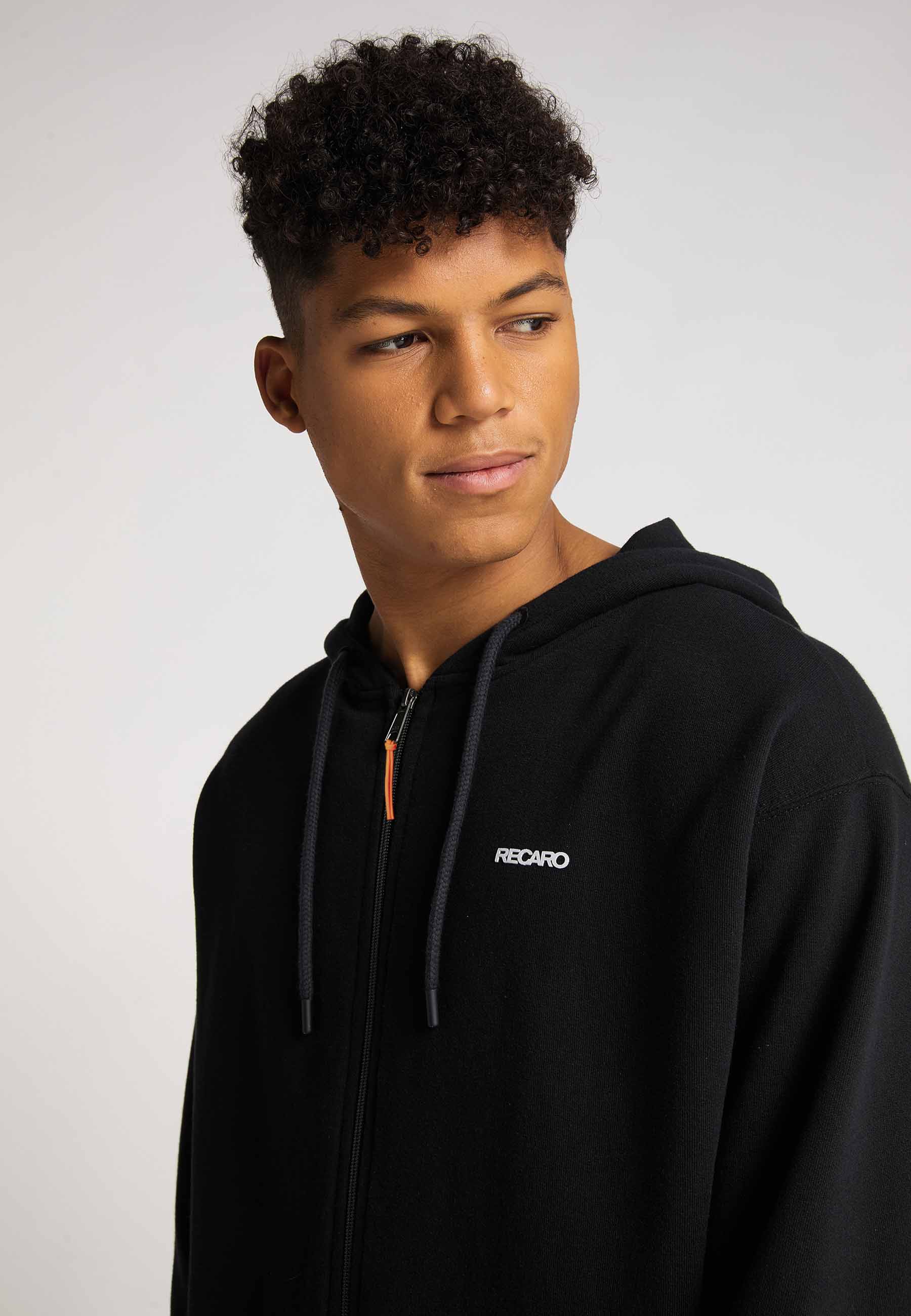Ein junger Mann mit kurzen lockigen Haaren trägt eine locker sitzende Hoodiejacke Originals von Recaro mit Bruststickerei und blickt vor einem schlichten hellen Hintergrund zur Seite.