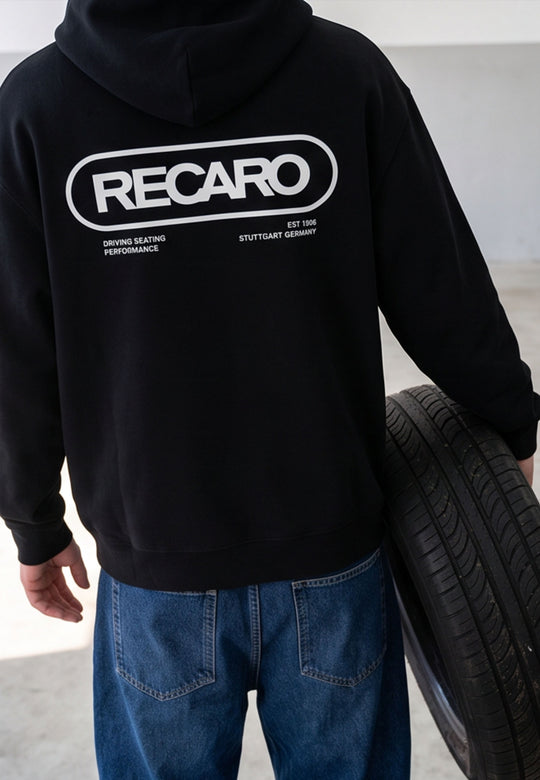 Eine Person in einer schwarzen RECARO Shop-Hoodiejacke im Retro-Stil mit Aufdruck auf dem Rücken und blauen Jeans steht in einem Raum und hält einen Autoreifen unter dem Arm – eine perfekte Verkörperung des „Racing-DNA“-Stils.