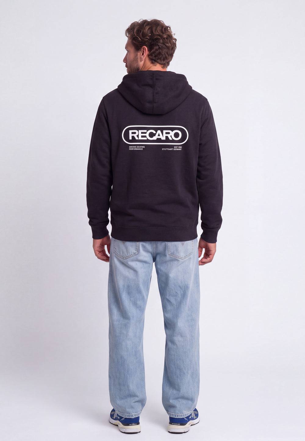 Ein Mann steht mit dem Rücken zur Kamera und trägt eine schwarze Hoodiejacke „Retro Backprint“ aus dem RECARO-Shop mit dem Aufdruck „RECARO“ auf dem Rücken, dazu hellblaue Jeans und blaue Sneaker vor einem schlichten weißen Hintergrund – ein Bild, das die Rennsport-DNA dieses charakteristischen RECARO-Modells perfekt einfängt.