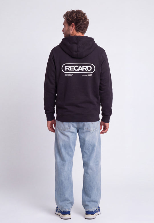 Ein Mann steht mit dem Rücken zur Kamera und trägt eine schwarze Hoodiejacke „Retro Backprint“ aus dem RECARO-Shop mit dem Aufdruck „RECARO“ auf dem Rücken, dazu hellblaue Jeans und blaue Sneaker vor einem schlichten weißen Hintergrund – ein Bild, das die Rennsport-DNA dieses charakteristischen RECARO-Modells perfekt einfängt.