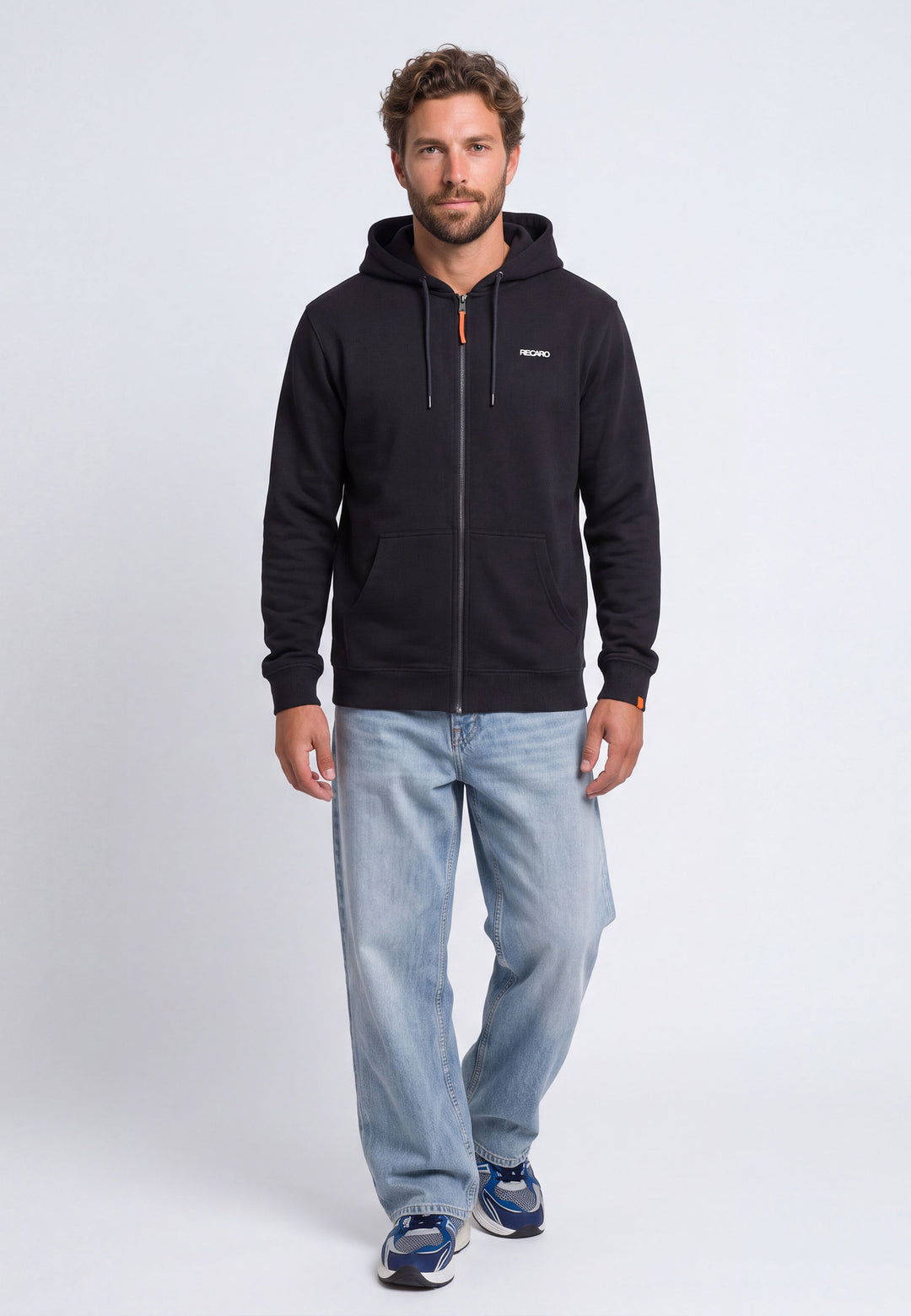 Ein Mann, der eine schwarze RECARO Shop-Hoodiejacke im Retro-Stil mit „Racing-DNA“-Aufdruck auf dem Rücken, hellblaue Jeans und blaue Sneaker trägt, steht mit dem Blick nach vorne vor einem schlichten weißen Hintergrund.