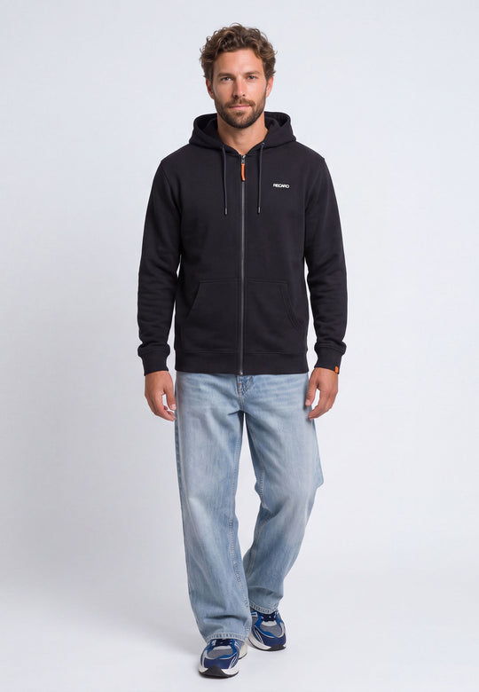 Ein Mann, der eine schwarze RECARO Shop-Hoodiejacke im Retro-Stil mit „Racing-DNA“-Aufdruck auf dem Rücken, hellblaue Jeans und blaue Sneaker trägt, steht mit dem Blick nach vorne vor einem schlichten weißen Hintergrund.