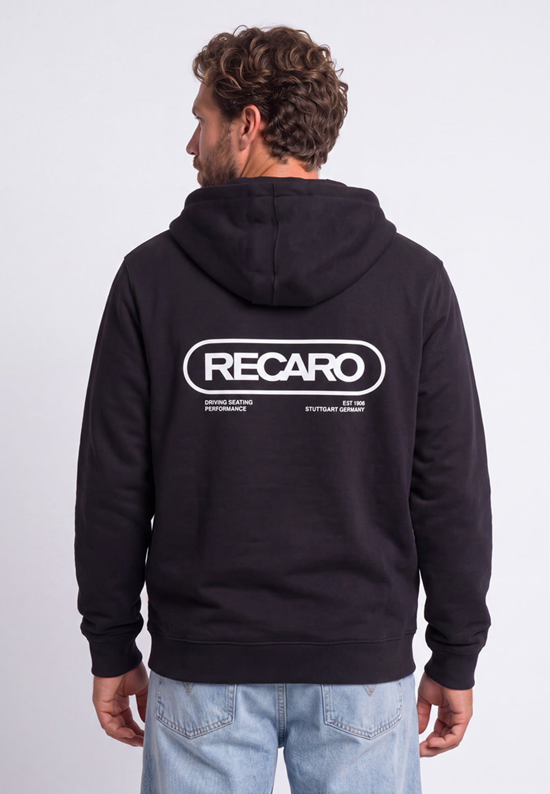 Ein Mann trägt eine schwarze Hoodiejacke „Retro Backprint“ aus dem RECARO Shop, auf deren Rücken das Wort „RECARO“ sowie weiterer weißer Text aufgedruckt sind. Er steht mit dem Rücken zur Kamera, wodurch das von der „Racing-DNA“ inspirierte Design dieses Motorsport-Hoodies besonders gut zur Geltung kommt.