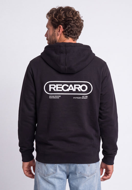 Ein Mann trägt eine schwarze Hoodiejacke „Retro Backprint“ aus dem RECARO Shop, auf deren Rücken das Wort „RECARO“ sowie weiterer weißer Text aufgedruckt sind. Er steht mit dem Rücken zur Kamera, wodurch das von der „Racing-DNA“ inspirierte Design dieses Motorsport-Hoodies besonders gut zur Geltung kommt.
