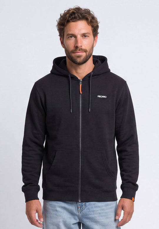 Ein Mann mit kurzen braunen Haaren und Bart trägt die schwarze RECARO Shop Hoodiejacke Retro Backprint, auf deren Brust „Racing-DNA“ aufgedruckt ist, dazu hellblaue Jeans, während er vor einem schlichten hellen Hintergrund steht.
