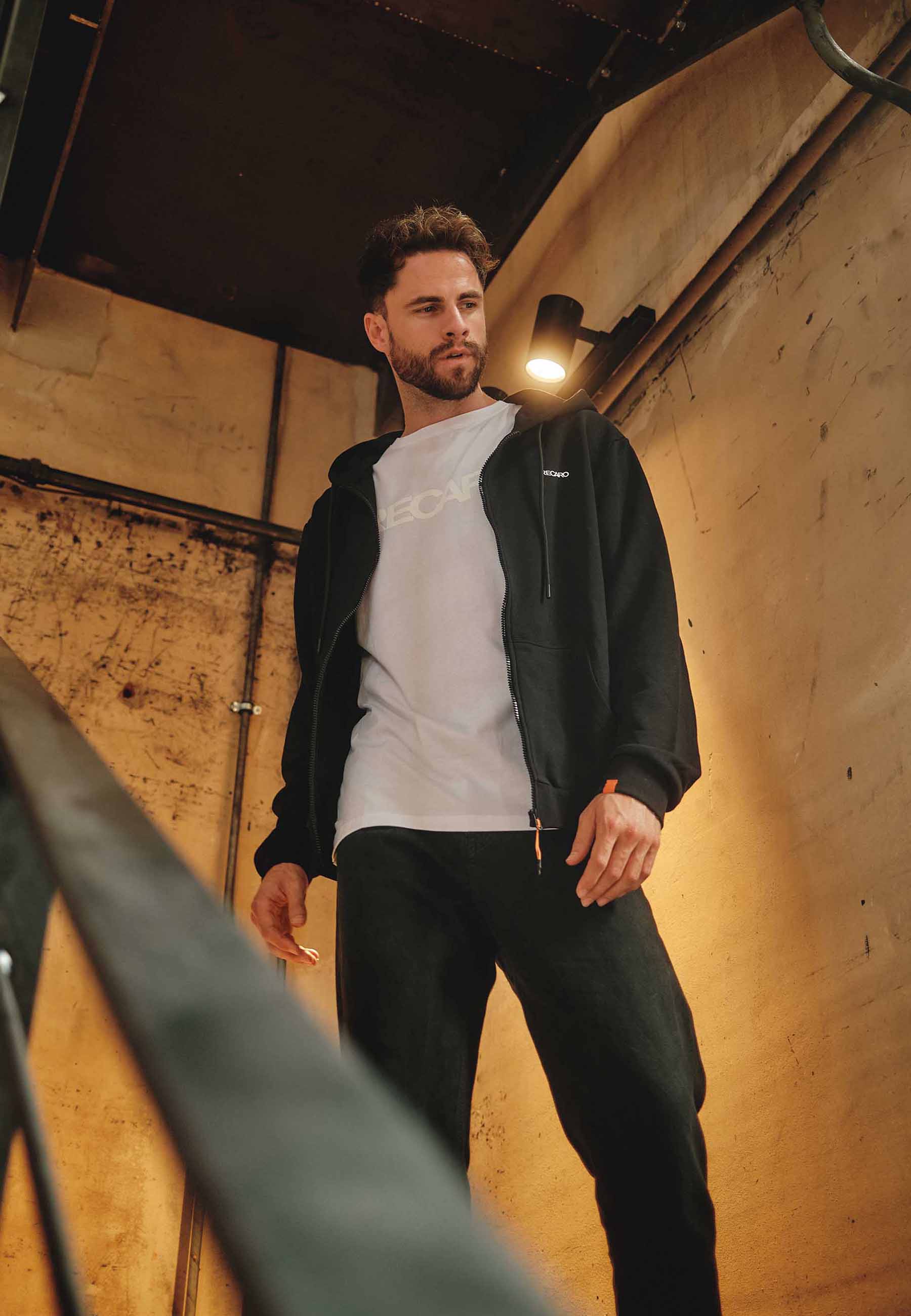 Ein Mann mit kurzen Haaren und Bart trägt ein weißes T-Shirt, eine schwarze Hose und die Unisex-Hoodiejacke Originals von Recaro_Merch_Daniel und steht auf einer Industrietreppe mit Betonwänden und warmer Beleuchtung für einen lässigen Look.