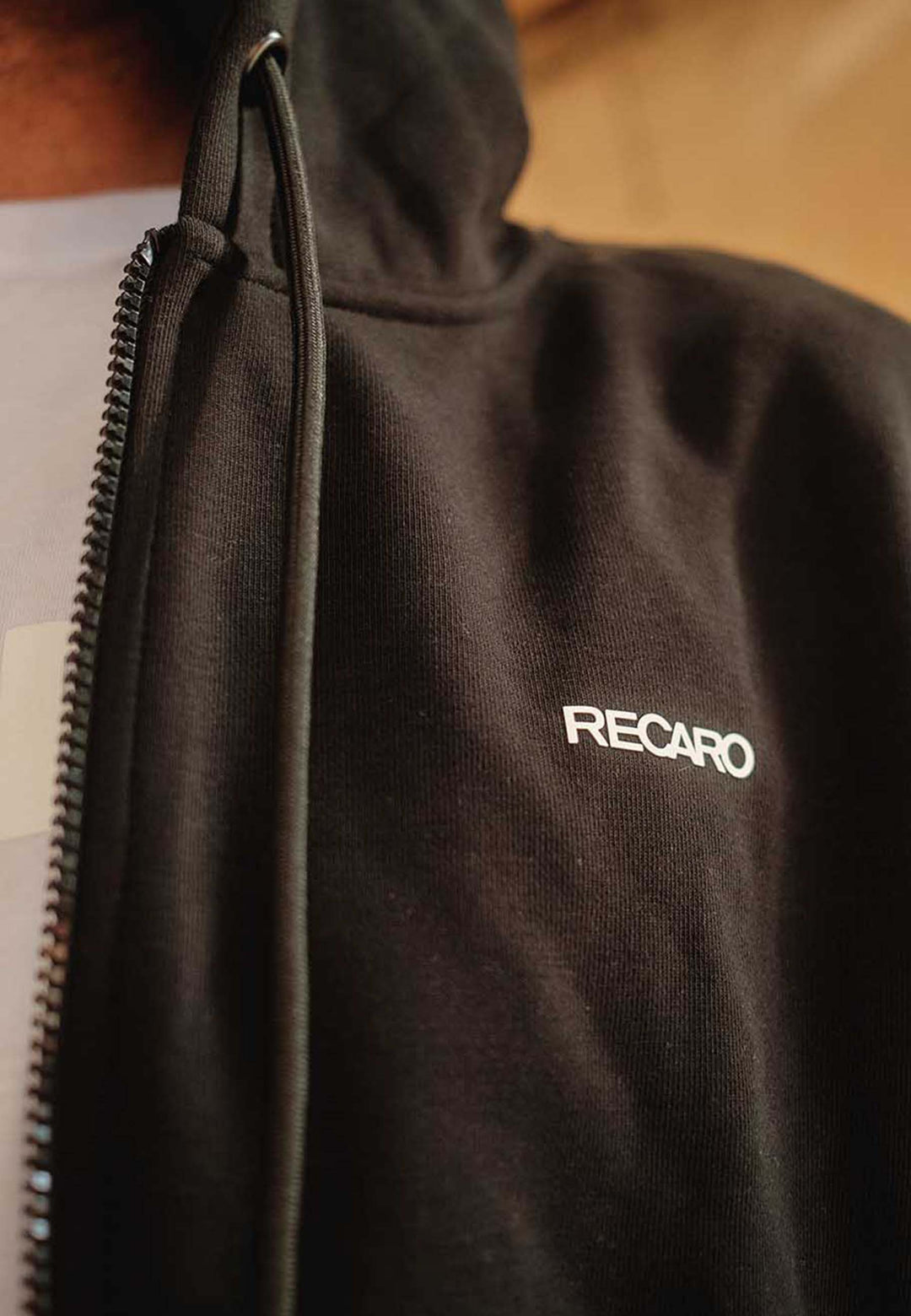 Nahaufnahme einer Person, die die schwarze Hoodiejacke Originals von Recaro_Merch_Daniel trägt, mit dem Schriftzug "RECARO" in Weiß auf der Brust und einer lockeren Unisex-Passform. Nur ein Teil des Nackens und der Schultern ist sichtbar, was für einen eleganten, subtilen Look sorgt.