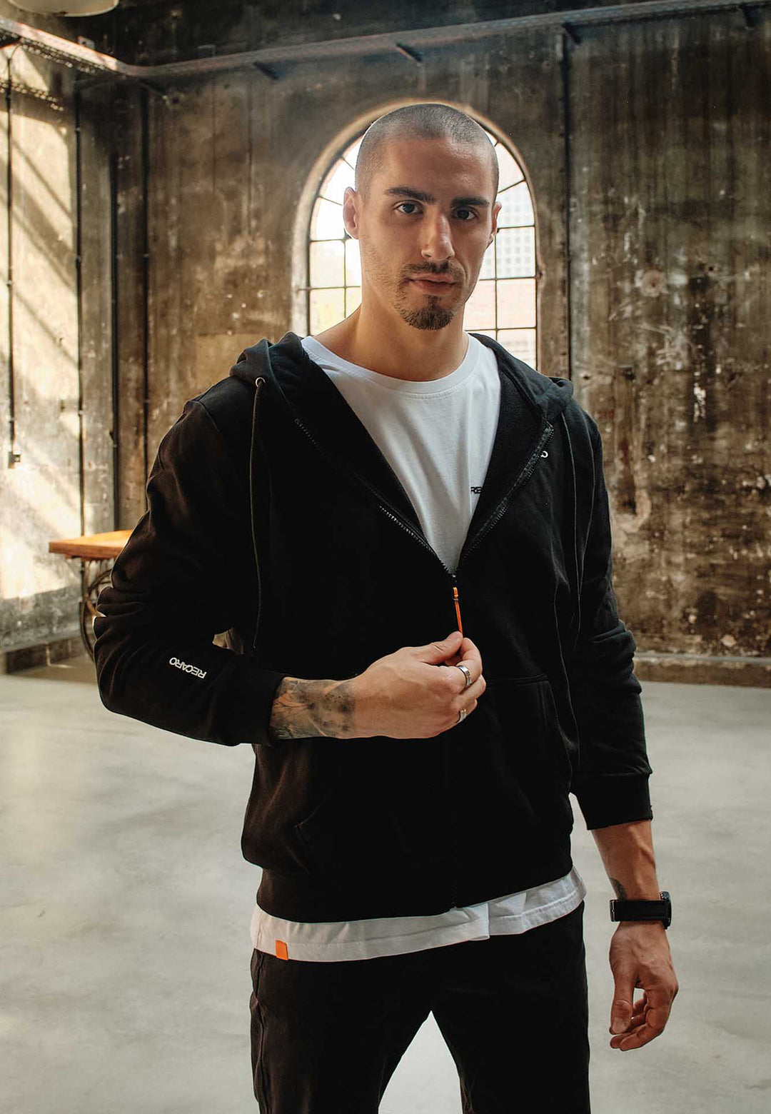 Ein Mann mit kurzen Haaren und gestutztem Bart steht an einem gewölbten Fenster in einem Innenraum, trägt eine Recaro_Merch_Daniel Hoodiejacke Originals über einem weißen T-Shirt und blickt mit einer Hand am Reißverschluss in die Kamera.