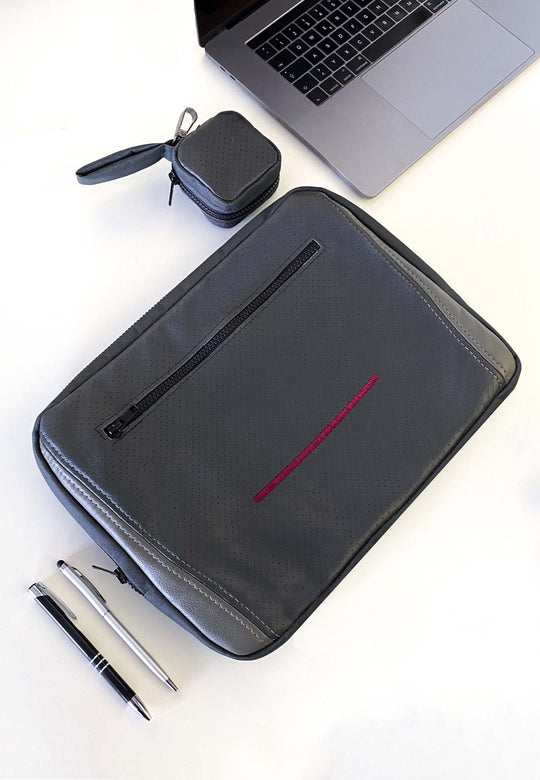 Die Tech & Travel Laptop Tasche aus der Upcycling Kollektion des RECARO Shops ist eine graue Hülle aus RECARO-Sitzleder mit einer Reißverschlusstasche vorne, einem roten Akzentstreifen, einem passenden Beutel und Stiften – geöffnet auf einer weißen Oberfläche dargestellt.