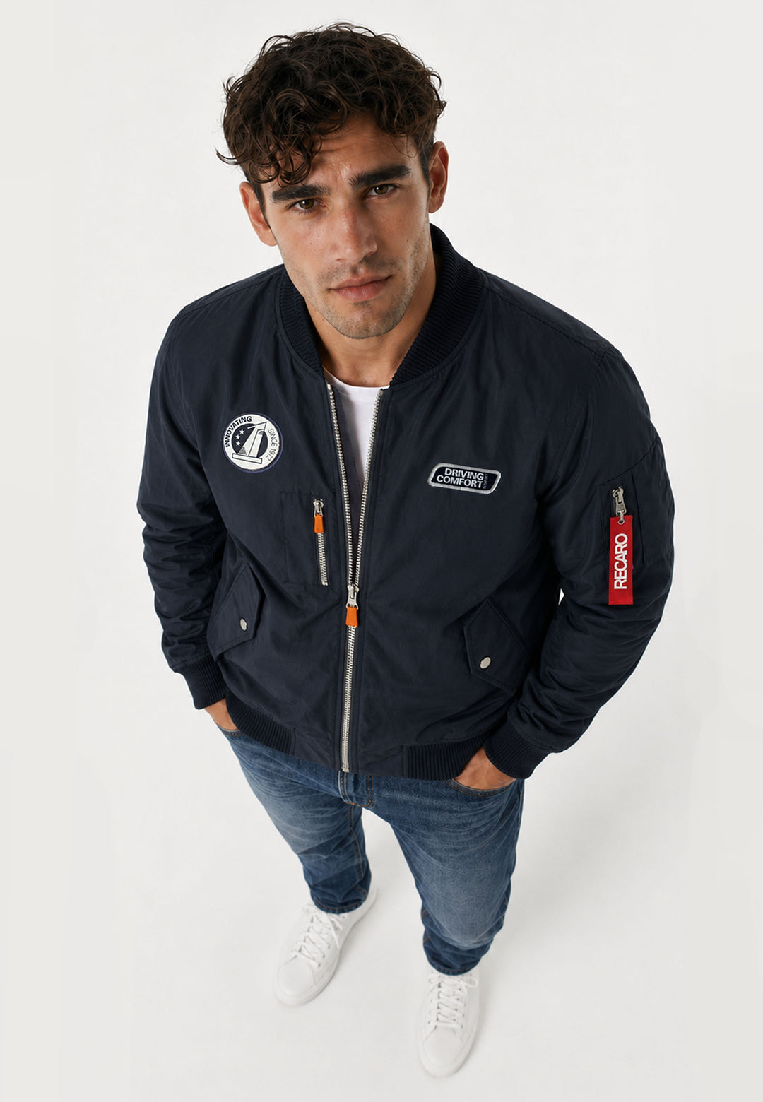 Ein Mann mit lockigen braunen Haaren trägt eine marineblaue Pilotenjacke RECARO von RECARO Shop, gepaart mit einem weißen T-Shirt, blauen Jeans und weißen Sneakers. Er steht mit den Händen in den Hosentaschen und blickt in die Kamera vor einem schlichten Hintergrund.