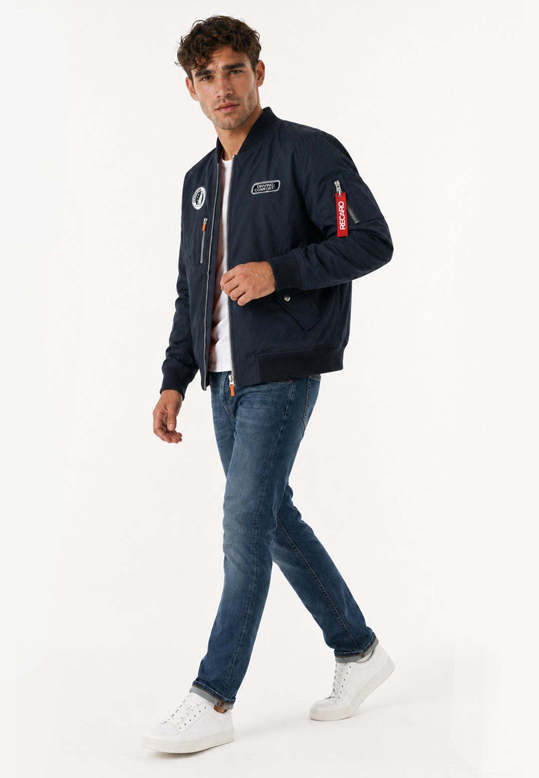 Ein Mann läuft selbstbewusst vor einem schlichten weißen Hintergrund. Er trägt eine navyfarbene Pilotenjacke RECARO aus dem RECARO Shop, gepaart mit blauen Jeans und weißen Sneakers.
