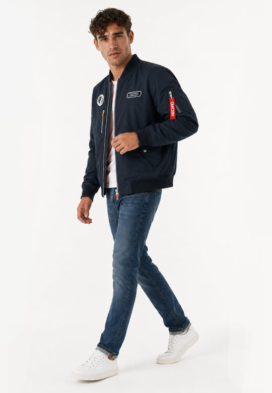 Ein Mann läuft selbstbewusst vor einem schlichten weißen Hintergrund. Er trägt eine navyfarbene Pilotenjacke RECARO aus dem RECARO Shop, gepaart mit blauen Jeans und weißen Sneakers.