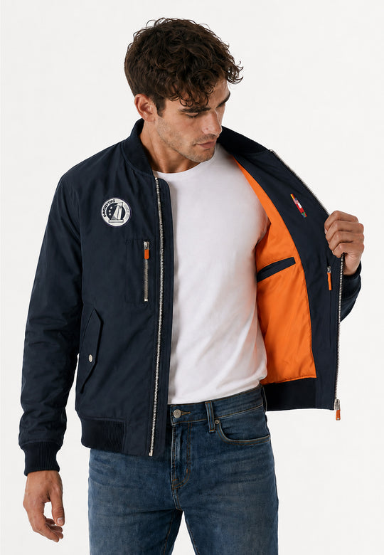 Ein Mann in weißem T-Shirt und Jeans modelliert die Pilotenjacke RECARO, navy aus dem RECARO Shop und hält sie auf, so dass das orangefarbene Innenfutter, die Innentasche und der Logoaufnäher auf der Brust sichtbar werden.
