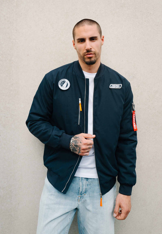 Ein Mann mit kurzen Haaren, einem gepflegten Bart und einem Tattoo auf der Hand trägt die marineblaue Pilotenjacke RECARO aus dem RECARO Shop über einem weißen T-Shirt und hellblauen Jeans und steht vor einer schlichten beigen Wand.