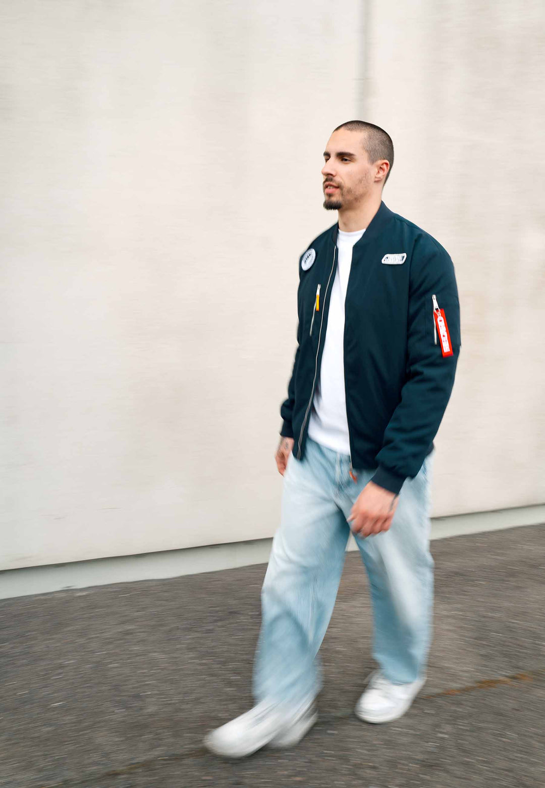 Ein Mann mit kurzen Haaren und Bart geht draußen auf einem Bürgersteig entlang. Er trägt die marineblaue Pilotenjacke RECARO aus dem RECARO Shop, dazu hellblaue Jeans und weiße Sneaker, vor dem Hintergrund einer schlichten, hellen Wand.