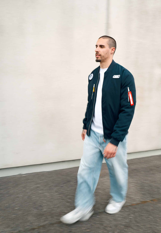 Ein Mann mit kurzen Haaren und Bart geht draußen auf einem Bürgersteig entlang. Er trägt die marineblaue Pilotenjacke RECARO aus dem RECARO Shop, dazu hellblaue Jeans und weiße Sneaker, vor dem Hintergrund einer schlichten, hellen Wand.