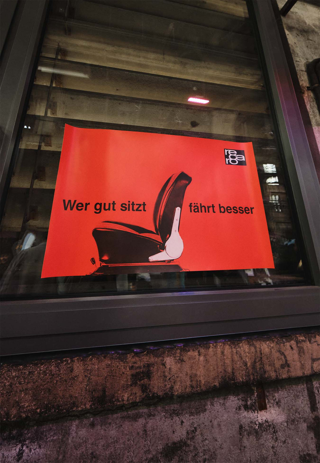 Das RECARO Shop Poster "Wer gut sitzt fährt besser" mit einer Autositzgrafik und deutschem Text ist an einem Fenster mit einer Holzkonstruktion und einer Betonwand im Hintergrund angebracht.