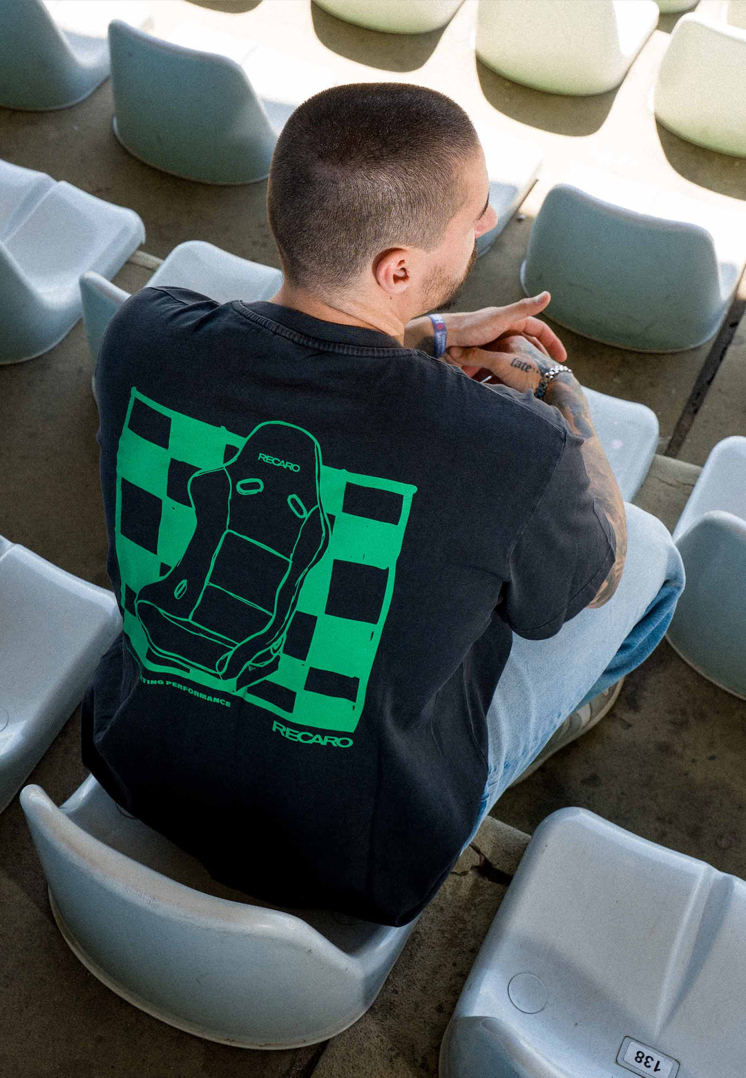 Eine Person mit kurzen Haaren sitzt auf weißen Stadion-Sitzen und trägt ein schwarzes RECARO Shop-T-Shirt mit der Aufschrift „Pole Position“ auf dem Rücken und dem Vermerk „Garment Washed“, das auf der Rückseite mit einer grünen Rennsitz-Grafik und einem Karomuster verziert ist – eine perfekte Mischung aus Motorsport- und Streetwear-Stil.