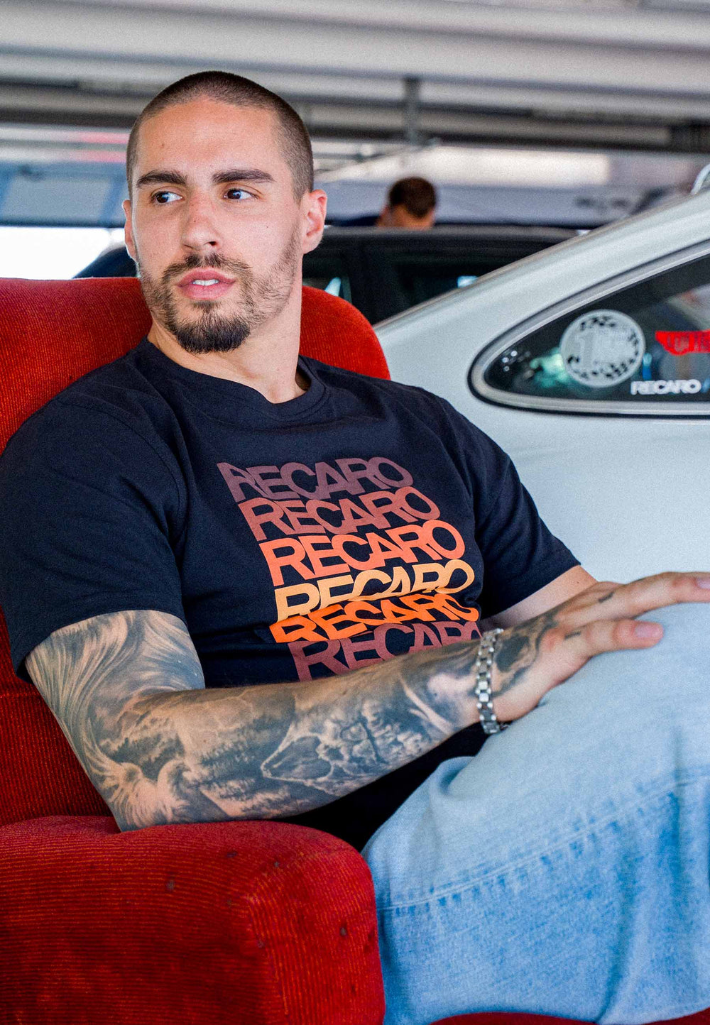 Ein Mann mit Tattoos sitzt auf einem roten Stuhl; er trägt das schwarze T-Shirt „Spektrum“ von Recaro_Merch_Daniel und hellblaue Jeans und strahlt vor dem Hintergrund eines weißen Autos einen Retro-Look aus.