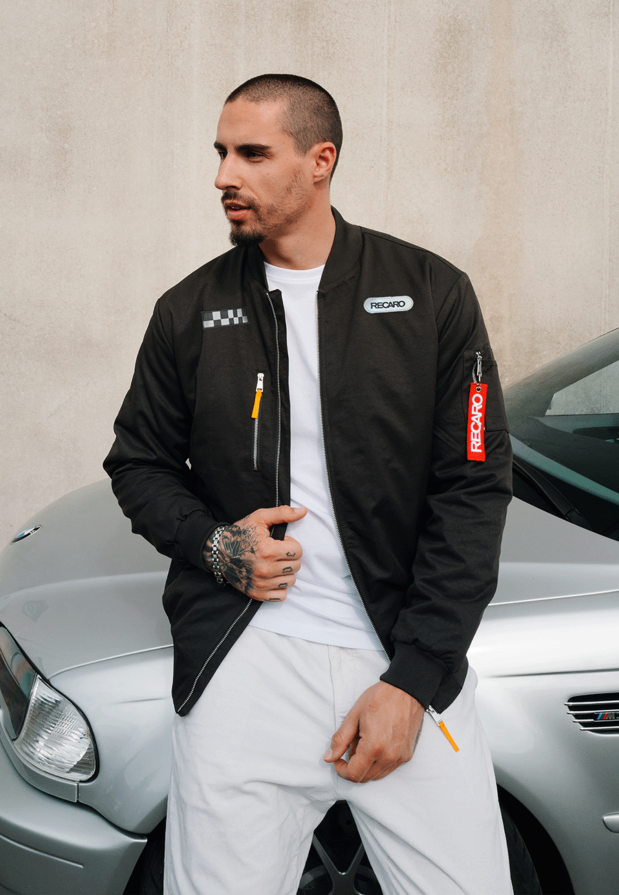 Ein Mann mit kurzen Haaren und Tattoos trägt die Racing Jacke RECARO in Anthrazit aus dem RECARO Shop, die im klassischen Bomber-Stil gehalten ist, dazu eine weiße Hose. Er steht neben einem silbernen Auto vor einer schlichten beigen Wand.