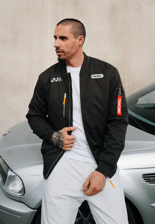 Ein Mann mit kurzen Haaren und Tattoos trägt die Racing Jacke RECARO in Anthrazit aus dem RECARO Shop, die im klassischen Bomber-Stil gehalten ist, dazu eine weiße Hose. Er steht neben einem silbernen Auto vor einer schlichten beigen Wand.