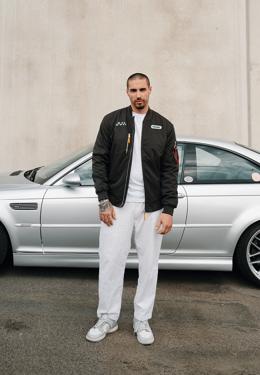 Ein Mann, der eine anthrazitfarbene RECARO-Rennjacke aus dem RECARO-Shop trägt – eine wind- und wasserabweisende Jacke im Bomber-Stil –, kombiniert diese mit einer weißen Hose, während er vor einem silbernen Auto steht, das neben einer beigen Wand geparkt ist.