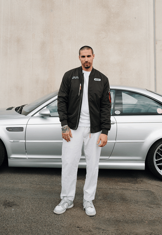 Ein Mann, der eine anthrazitfarbene RECARO-Rennjacke aus dem RECARO-Shop trägt – eine wind- und wasserabweisende Jacke im Bomber-Stil –, kombiniert diese mit einer weißen Hose, während er vor einem silbernen Auto steht, das neben einer beigen Wand geparkt ist.