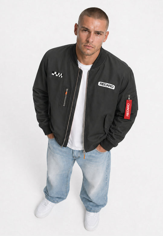 Ein Mann mit kurzen Haaren präsentiert die anthrazitfarbene Racing Jacke RECARO aus dem RECARO Shop, die in einer klassischen Bomber-Silhouette mit markanten RECARO-Aufnähern gestaltet ist. Er kombiniert die wind- und wasserabweisende Jacke mit einem weißen T-Shirt, hellblauen Jeans und weißen Sneakern. Vor einem weißen Hintergrund blickt er in die Kamera und lässt dabei die auffällige Jacke im Mittelpunkt stehen.