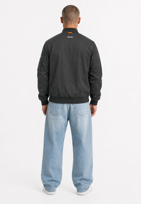Eine Person steht mit dem Rücken zur Kamera und trägt die „Racing Jacke RECARO“ in Anthrazit aus dem RECARO Shop, die sich durch einen Schriftzug auf der Rückseite und eine klassische Bomber-Silhouette auszeichnet. Die Jacke ist wind- und wasserabweisend und wird vor einem schlichten weißen Hintergrund mit einer lockeren hellblauen Jeans und weißen Schuhen kombiniert.