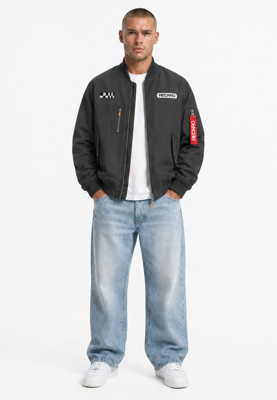 Ein Mann blickt vor einem schlichten weißen Hintergrund in die Kamera. Er trägt eine anthrazitfarbene RECARO-Rennjacke aus dem RECARO Shop in klassischer Bomber-Silhouette mit Rennsport-Aufnähern, dazu ein weißes T-Shirt, weite hellblaue Jeans und weiße Sneaker.
